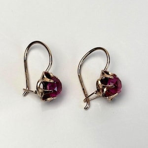 Pode incluir: Um par de brincos com pedras redondas facetadas em rosa profundo, fixadas em garras de metal dourado. Cada brinco possui um fecho de gancho curvo que se estende da base da cravação, criando um design pendente clássico com acabamento polido.