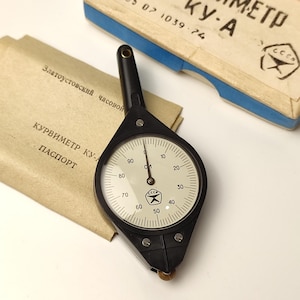 May include: A black and white analogue gauge with a needle pointing to 0. The gauge is labelled "КУРВИМЕТР КУ-А" and "ТУ 25 07 1039-74". The gauge is attached to a black plastic handle.