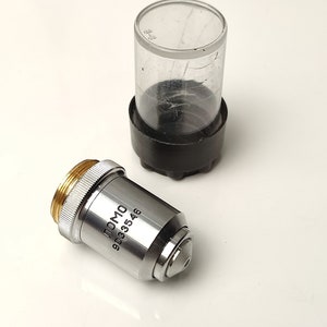 May include: A silver microscope objective lens with the text "LOMO 9033546" printed on the side. The lens is sitting on a white surface next to a clear plastic container with a black lid.
