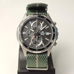 May include: A silver chronograph wristwatch with a black face and a green and white striped fabric strap. The watch face has a date window, three subdials, and the text "EDIFICE CASIO WR 100M".