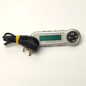 Könnte beinhalten: Ein silberner Kenwood MP3-Player mit einem schwarzen Kabel und einem gelben Gummiband. Der Player hat einen grünen Bildschirm und Steuertasten. Das Kenwood-Logo ist auf dem Bildschirm zu sehen.