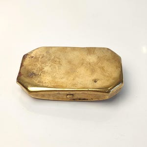 May include: A rectangular brass case with beveled edges and a polished, metallic gold-toned finish. The surface shows subtle signs of wear and texture, with a small latch visible on the side of the compact, hinged metal container.
