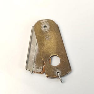 May include: A vintage brass-toned metal razor blade holder with a rectangular shape, featuring a circular cutout, a small metal loop at the bottom, and a silver-toned blade partially extended from the side. The surface shows signs of wear.