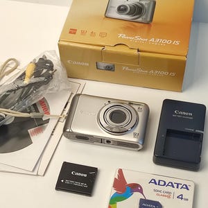 May include: A silver Canon PowerShot A3100 IS digital camera with its original yellow box, a black battery charger, a Canon NB-8L battery pack, a 4GB ADATA SDHC memory card in its packaging, a wrist strap, and various cables and manuals.