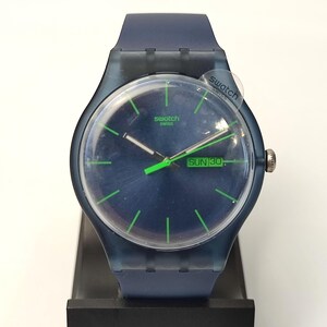 Puede incluir: Un reloj de pulsera Swatch con caja azul oscuro translúcida y correa de silicona a juego. La esfera azul oscuro presenta marcadores de hora en verde brillante, manecillas verdes a juego y una ventana de día y fecha que muestra SUN 30. El logo Swatch está impreso.