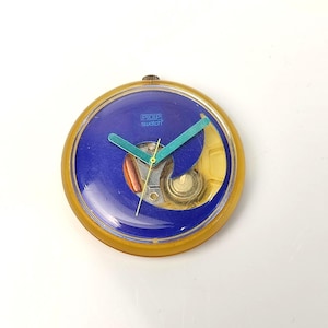May include: A vintage Swatch Pop watch with a blue face and teal hands. The watch has a transparent yellow case and the text "POP swatch" is visible on the face. The watch is round and has a gold-colored crown.