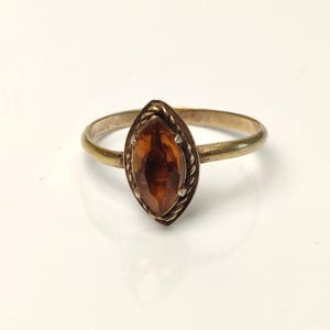 Peut inclure: Une bague vintage avec une monture dorée et une pierre de couleur ambre en forme de marquise. La pierre est sertie dans une lunette argentée avec un motif de corde torsadée. La bague est présentée sur une surface blanche.