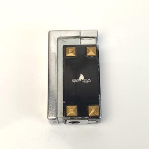May include: A silver and black lighter with gold-coloured square studs. The lighter has the brand name "BAFA" printed on the black surface, along with a flame symbol. The lighter is rectangular and has a hinged top.