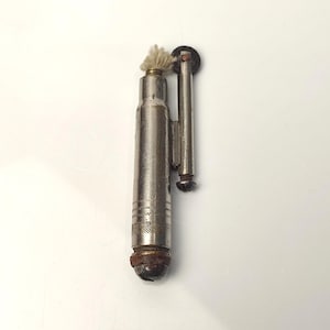 Puede incluir: Un encendedor plateado vintage con un cuerpo cilíndrico y un brazo articulado. El encendedor tiene una mecha en la parte superior y un patrón texturizado cerca de la parte inferior. La base del encendedor muestra signos de edad y desgaste.