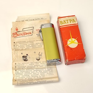 May include: A vintage lighter, a box, and instructions are displayed. The lighter is olive green with a silver top. The box is red with a yellow starburst design. The instructions are in a foreign language.