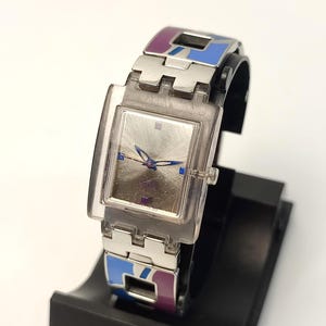 Könnte beinhalten: Eine silberne Armbanduhr mit rechteckigem Zifferblatt und transparentem Kunststoffarmband. Das Armband hat blaue und violette Akzente. Die Uhr wird auf einem schwarzen Ständer vor weißem Hintergrund präsentiert.