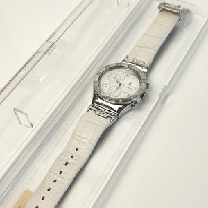 May include: A silver-tone chronograph wristwatch featuring a white dial with three sub-dials and a white leather strap with a textured crocodile-embossed pattern. The watch is housed in a clear plastic rectangular display case with the word Swatch.