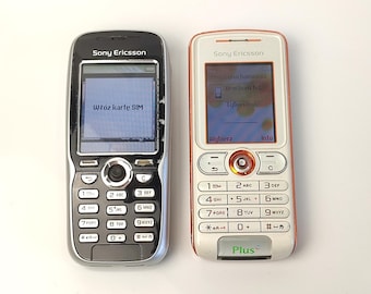 May include: Two vintage Sony Ericsson cell phones. One is black and silver, displaying "Włóz kartę SIM" on the screen. The other is white and orange, with a screen showing menu options. Both phones have physical keypads.