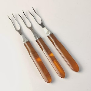Peut inclure: Trois fourchettes de service vintage avec des dents en acier inoxydable et des manches couleur ambre. Les fourchettes sont disposées en diagonale, mettant en valeur leur design élégant. Les manches ont une finition lisse et brillante.