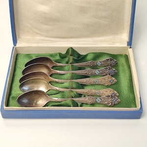 May include: Set of six antique silver spoons in a blue box with a green satin lining. The spoons have ornate handles with floral and turquoise enamel details. A vintage silverware set.