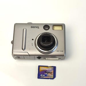 May include: A silver BenQ DC C40 digital camera with a 3x optical zoom lens and a flash. Below the camera is a blue Ultra SD memory card with 256MB of storage.