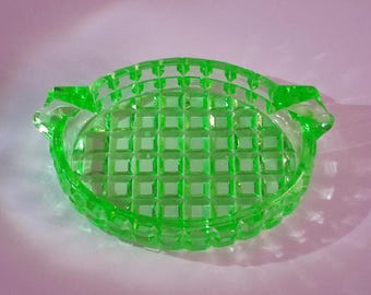 May include: A vibrant green glass ashtray with a grid pattern and two small handles. The ashtray is oval-shaped and sits on a light purple surface. The glass has a slight glow, suggesting it may be vintage or made with a special glass.