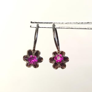 Op de afbeelding: Een paar zilverkleurige oorhangers. Elke oorbel heeft een bloemvormig ontwerp met een roze edelsteen in het midden. De oorbellen zijn opgehangen aan een gebogen haak.