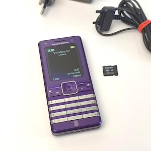 Op de afbeelding: Een paarse Sony Ericsson mobiele telefoon met een scherm met "Vodafone UA Lebara" en de tijd. Inclusief een oplader, een geheugenkaart en een zwarte voedingsadapter. De telefoon heeft een toetsenbord met cijfers en symbolen.