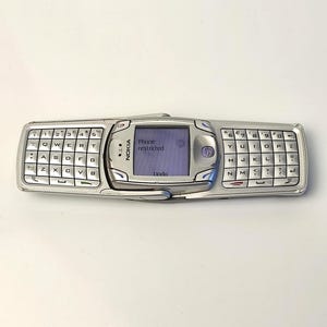 Puede incluir: Un teléfono móvil Nokia plateado en orientación horizontal con un teclado QWERTY completo dividido a ambos lados de una pantalla central. La pantalla muestra el texto Phone restricted y Undo en una fuente azul oscuro sobre un fondo claro.