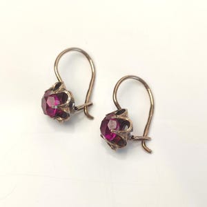 May include: A pair of earrings featuring round, faceted, deep pink stones set in metallic, pronged settings. Each earring has a curved, hinged wire hook for attachment, with the metal showing a slightly darkened, aged finish on the prongs.
