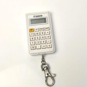 Puede incluir: Una calculadora Canon blanca con una pantalla digital que muestra "0". La calculadora tiene un accesorio para llavero. Tiene un teclado estándar con números y símbolos matemáticos.