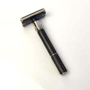 Puede incluir: Una maquinilla de afeitar de seguridad negra y plateada con un mango acanalado. El cabezal de la maquinilla es plateado y el mango es negro con detalles plateados. La maquinilla está diseñada para afeitarse y tiene un aspecto clásico y vintage.