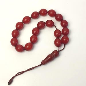 May include: A string of prayer beads featuring oval, reddish-brown beads. The beads are connected by a matching brown cord, with a decorative, elongated bead at the end. The background is plain white.