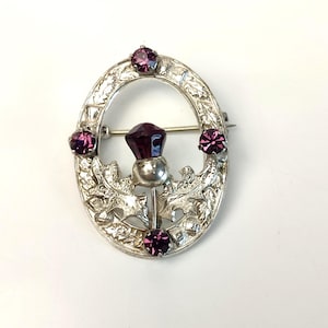 May include: An oval silver-toned brooch featuring a central thistle motif. The frame is decorated with embossed leaf patterns and four round, faceted purple crystals set at the top, bottom, and sides. A pin mechanism is visible on the back.