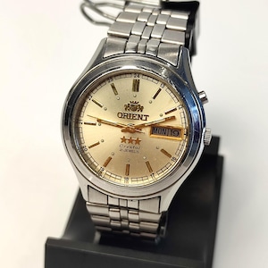May include: An Orient wristwatch with a silver-tone stainless steel link bracelet and a round case. The watch features a champagne-colored dial with gold-tone hour markers, hands, the Orient logo, three stars, and the text Crystal 21 JEWELS.
