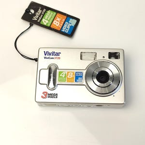 May include: Silver Vivitar ViviCam 3720 digital camera with a black tag. The camera has a lens, flash, and buttons. The tag reads "4 Mega Pixels Interpolation", "8x Zoom", and "Video Clips". The camera has "3 Mega Pixels" printed on it.
