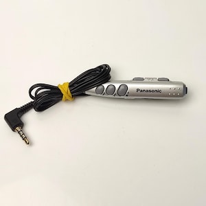 Puede incluir: Mando a distancia plateado de Panasonic con cable negro y conector jack de 3,5 mm. El mando tiene botones de volumen y control. Una goma elástica amarilla asegura el cable.