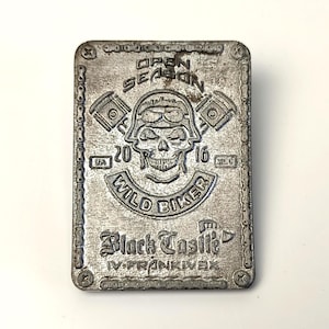 May include: A rectangular, silver-colored metal plaque with a biker theme. The plaque features a skull wearing a helmet, pistons, and the words "OPEN SEASON," "WILD BIKER," and "Black Castille." The year "2016" is also visible.