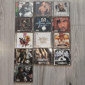 Puede incluir: Una colección de trece CD de música hip-hop dispuestos en una cuadrícula. Los álbumes presentan a varios artistas, incluidos 2Pac, Ludacris, Method Man, Ja Rule y Jay-Z, con portadas que muestran retratos, ilustraciones gráficas y etiquetas explícitas.