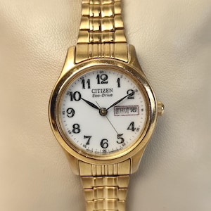May include: A gold-toned Citizen Eco-Drive wristwatch with a white face. The watch features black numerals, a date display, and a gold-toned link bracelet. The time reads Thursday the 16th.