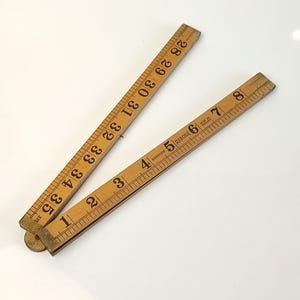 Puede incluir: Una regla plegable de madera vintage, con marcas en centímetros de 1 a 89. La regla es de madera marrón claro con marcas marrones oscuras. La marca "RABONE" es visible. La regla está parcialmente abierta, mostrando su diseño articulado.