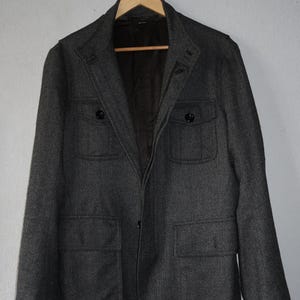 May include: A charcoal gray blazer with a subtle herringbone pattern. The jacket features two chest pockets, two lower pockets, and black button closures. The blazer is hanging on a wooden hanger against a neutral background.