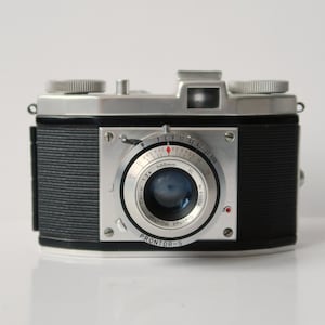 Puede incluir: Cámara vintage en blanco y negro con una lente prominente y diales. La cámara tiene un cuerpo rectangular con un agarre texturizado y una parte superior plateada. La lente está rodeada por un anillo plateado con marcas. Un equipo fotográfico clásico.