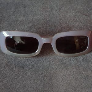 May include: Pale lavender sunglasses with dark lenses. The rectangular frames have a subtle, pearlescent sheen. The sunglasses are set against a gray background.