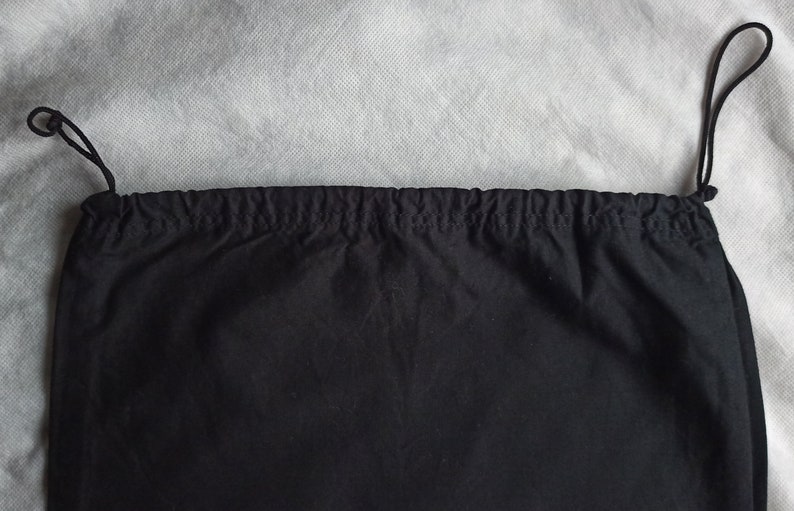 May include: A black drawstring bag with a gathered top and black cord ties. The bag is made of a dark fabric and is set against a light gray background. The drawstring cords are looped at the top.