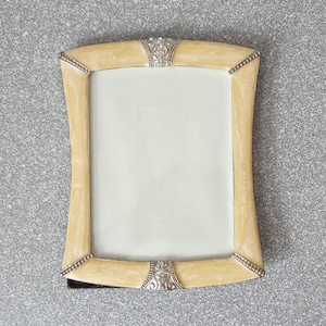 May include: A rectangular picture frame featuring a cream-colored, marbled enamel border with a glossy finish. The frame is accented with silver-toned metal corners and decorative center pieces at the top and bottom, each containing small clear crystals.
