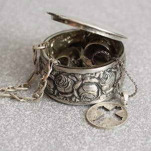 Op de afbeelding: Een ronde zilveren sieradendoos met een scharnierend deksel voorzien van een reliëfpatroon van rozen aan de buitenkant. De open doos bevat diverse zilveren sieraden, waaronder een dunne ketting met een ronde hanger met een kruisvormige uitsparing.