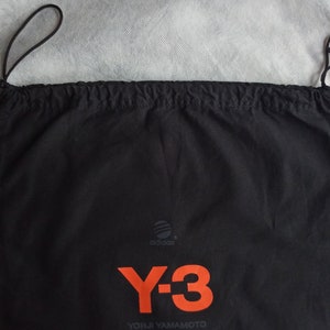 May include: Black drawstring bag with the Adidas logo and the Y-3 logo in orange. The text "YOHJI YAMAMOTO" is printed below the logo. The bag is made of a soft material and has a drawstring closure.