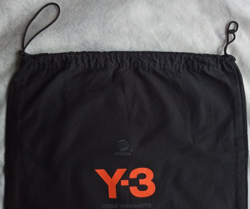 May include: Black drawstring bag with the Adidas logo and the Y-3 logo in orange. The text "YOHJI YAMAMOTO" is printed below the logo. The bag is made of a soft material and has a drawstring closure.