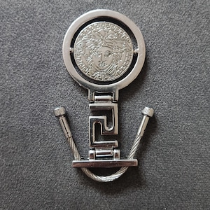 May include: Silver-toned keychain featuring a circular medallion with a Medusa head design. The keychain has a rectangular link and two cable-style attachments. The background is a dark gray surface.