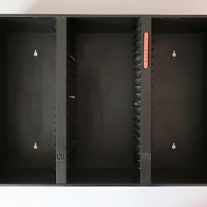 May include: A black plastic media storage rack with three vertical compartments designed for organizing discs. The unit features molded slots along the interior walls, keyhole mounting holes on the back panels, and the text "CD" and "LASERDISC" printed on the dividers.