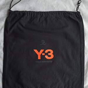 May include: Black drawstring bag with the Adidas logo and the Y-3 logo in orange. The text "YOHJI YAMAMOTO" is printed below the Y-3 logo. The bag has a drawstring closure.