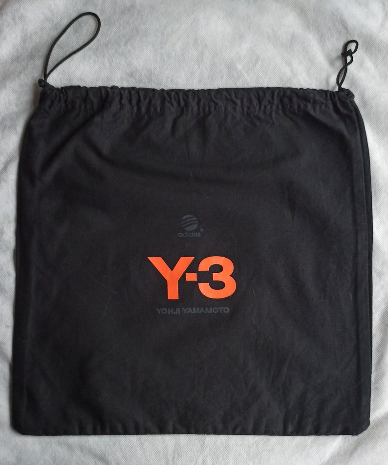 May include: Black drawstring bag with the Adidas logo and the Y-3 logo in orange. The text "YOHJI YAMAMOTO" is printed below the Y-3 logo. The bag has a drawstring closure.