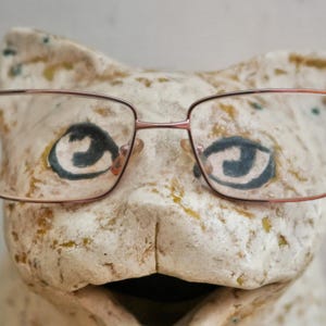 May include: A pair of rectangular eyeglasses with thin, metallic rose-gold frames positioned over the face of a textured, off-white ceramic cat figurine featuring hand-painted dark eyes with a swirling pattern and subtle yellow-green accents.