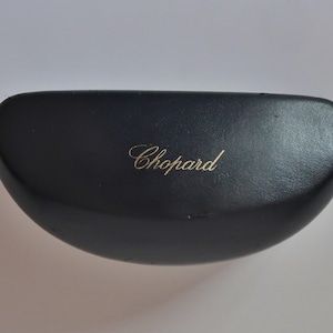 May include: A black, semi-circular hard-shell glasses case with a smooth, matte finish. The brand name Chopard is printed in elegant, gold-colored cursive script in the center of the top lid. The case features a curved, rounded design.